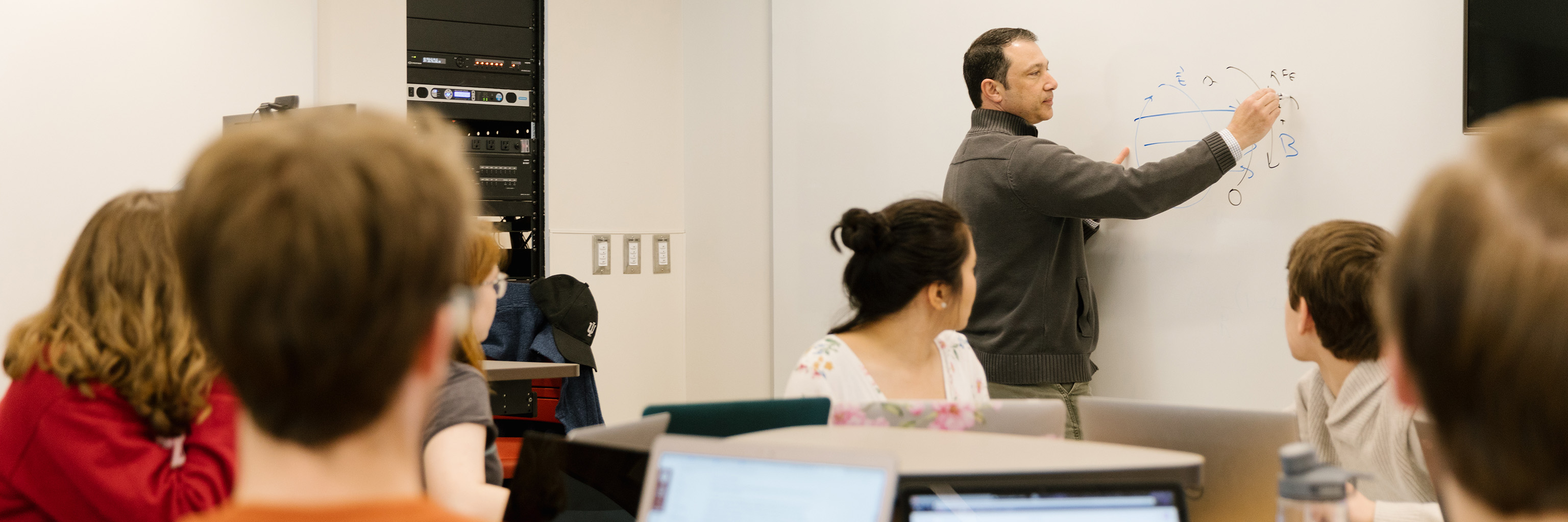 A professor illustrates a problem on a whiteboard in front of a group of students.